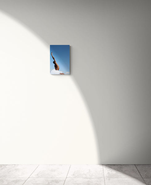 Black male dancing in the nature. His hand is oriented up onto the blue sky. We see him from behind. his armpit is glowing into the sunlight. Art print on a white wall.
